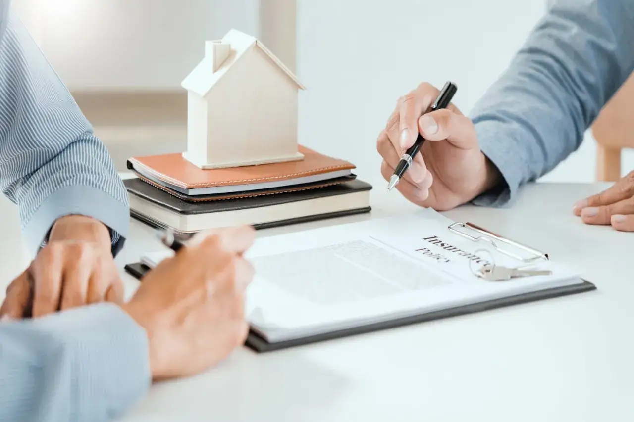 Deux mains signent un contrat d'assurance, une maison miniature sur des livres. Une clause tontine pourrait &ecirc;tre incluse.