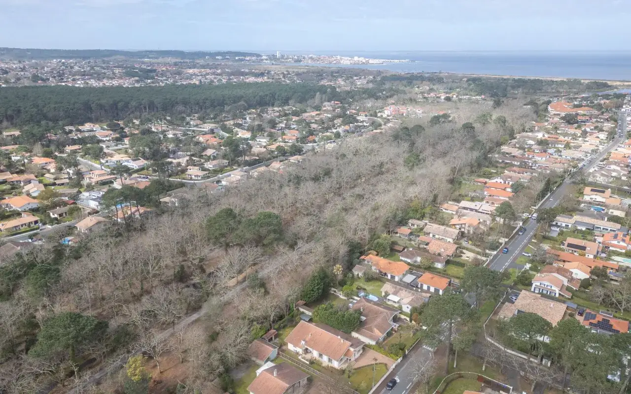 Vue aérienne de Gujan-Mestras, montrant un quartier résidentiel avec des maisons aux toits de tuiles, entouré de pins et bordé par l'océan au loin. L'urbanisme s'intègre à la nature.