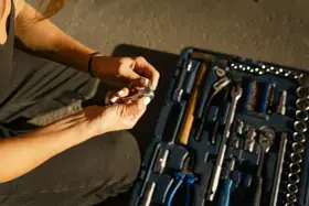 Mains féminines avec ongles blancs, manipulant une clé à cliquet. Une boîte à outils bleue est ouverte à côté, remplie d'outils. Une scène de travaux sans accord.