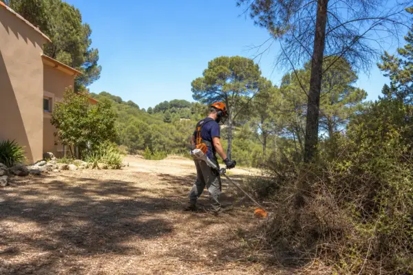 Un homme équipé pour le débroussaillement travaille près d'une maison, dans un paysage boisé. La jurisprudence débroussaillement est essentielle pour la prévention des incendies.